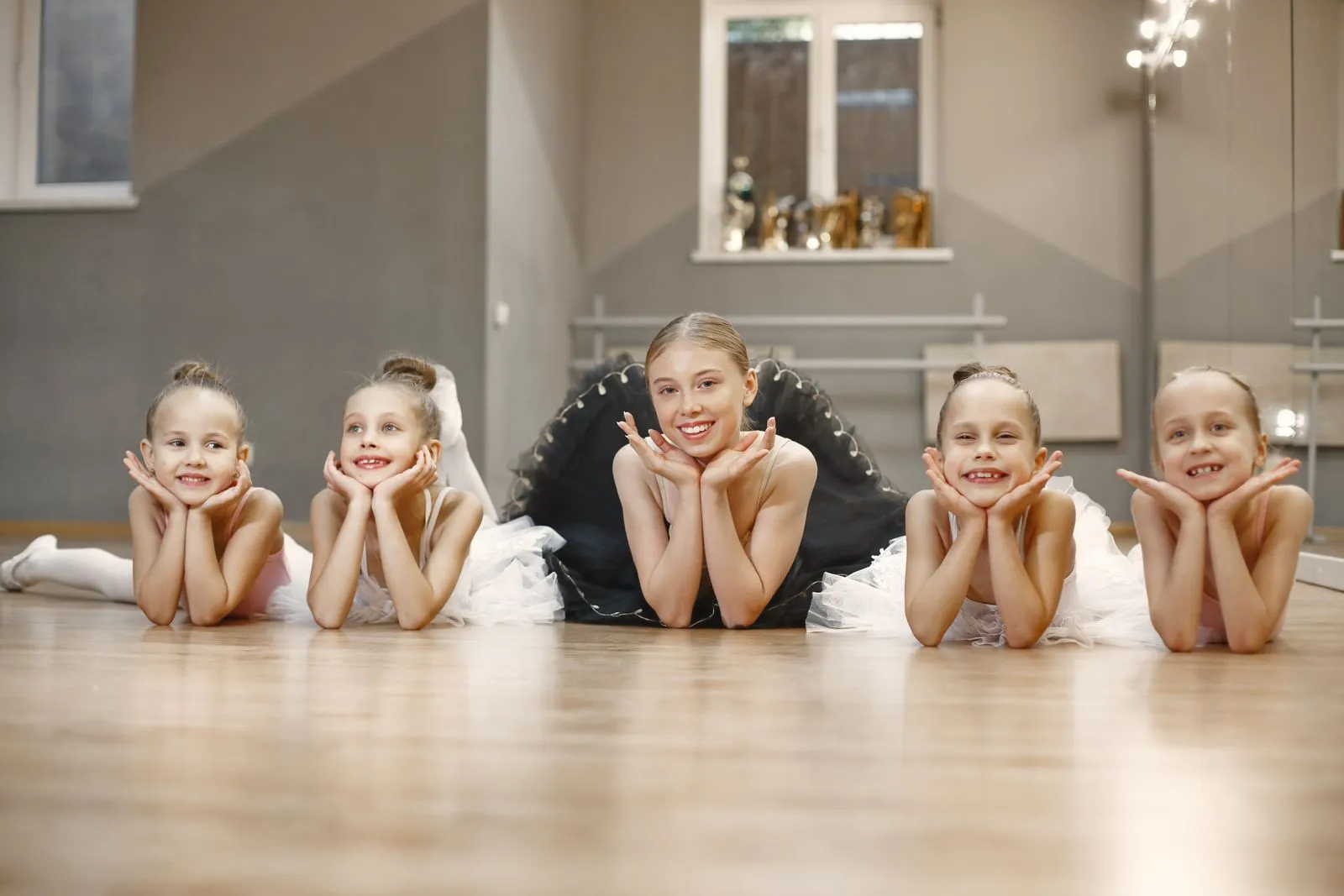 Five young ballerinas lying on the studio floor, smiling at the camera in tutus.